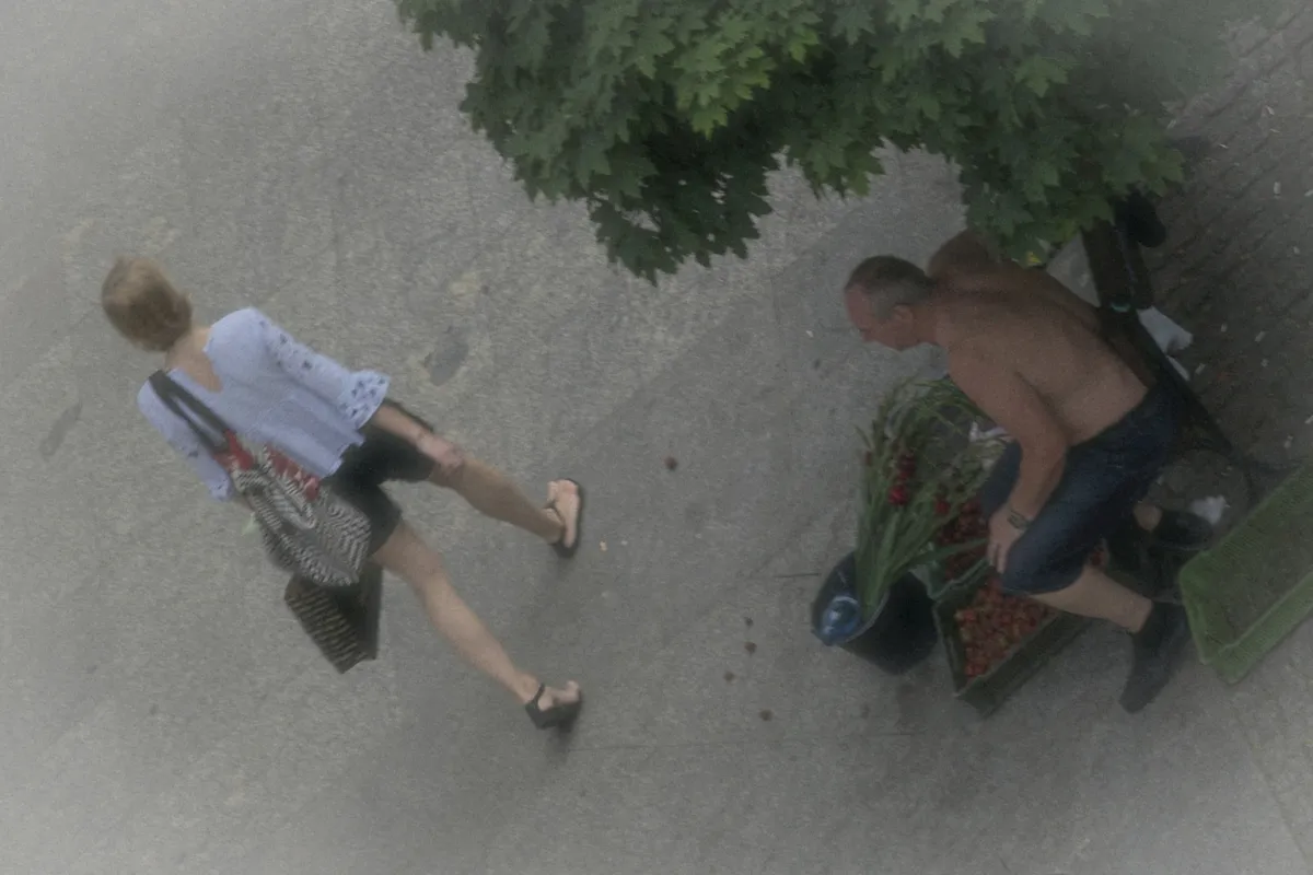 Overhead view of a woman walking past a shirtless street vendor seated under a tree
